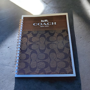 Puede incluir: Un cuaderno con espiral con una cubierta de cuero marrón con el logotipo de Coach y su patrón característico. La sección superior es de color marrón liso, mientras que la sección inferior muestra un patrón de 'C' repetido en un tono más oscuro de marrón. El cuaderno tiene un borde blanco.