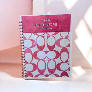 Può includere: Un taccuino a spirale con un motivo rosa e bianco. La sezione superiore è rosa pieno con il logo Coach in argento. Il taccuino è aperto, rivelando le pagine a motivi. Il design presenta il logo Coach.