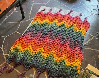 May include: A handmade, colorful crochet blanket with a chevron pattern. The blanket features a vibrant spectrum of colors, including orange, yellow, green, and purple. It has a scalloped edge with purple tassels, adding a decorative touch. The blanket is approximately 60 inches long.