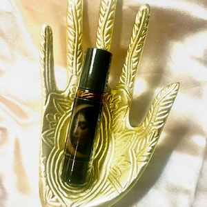 May include: A dark brown essential oil roller bottle with a black cap, featuring a woman's face with a crescent moon design. The bottle rests on a gold-colored hand-shaped decorative tray. The tray has intricate leaf-like patterns.