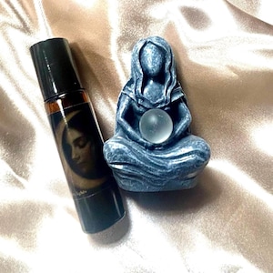 May include: A dark roller bottle with a woman's face on the label, next to a blue-grey statue of a seated figure holding a clear crystal ball. The items are on a satin surface.