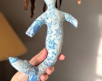 May include: A handmade mermaid doll with a blue and white patterned fabric body and a brown felt hair. The doll has outstretched arms and a fishtail. The doll is approximately 10 inches tall.