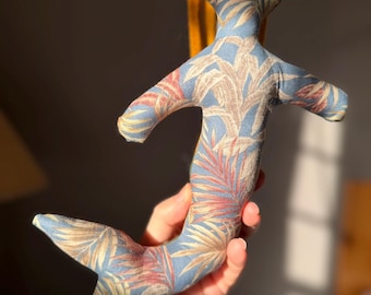 May include: A handmade mermaid doll with a blue fabric body featuring a tropical leaf pattern in shades of pink, beige, and yellow. The doll has a yellow hair and a fishtail. The doll is soft and plush.