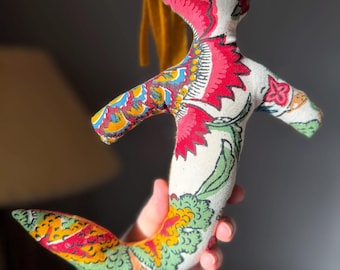 May include: A handmade mermaid doll with a cream-colored body and a colorful floral pattern. The doll has a red face, yellow hair, and a patterned tail with green, orange, and yellow accents. The doll is held in a hand.