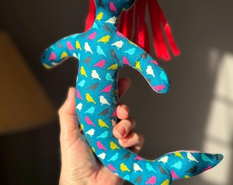 May include: A handmade mermaid doll with a blue body patterned with colorful birds. The doll has red hair and a fishtail. The doll is being held up in front of a window.