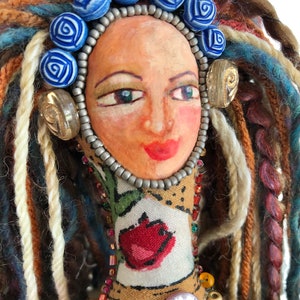 Detail of handpainted doll's face, grey, red, and gold beads and shell buttons, blue rose beads and multi-colored yarn hair