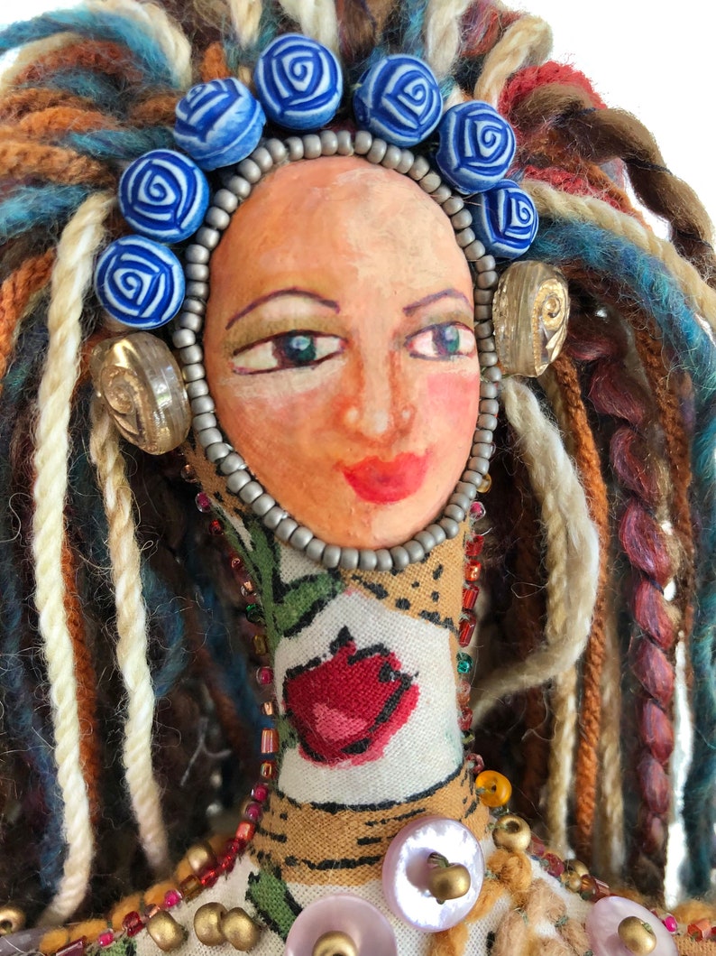 Detail of handpainted doll's face, grey, red, and gold beads and shell buttons, blue rose beads and multi-colored yarn hair