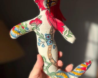 May include: A handmade fabric mermaid doll with a red head and tail. The doll's body is cream-colored and adorned with a colorful floral pattern in red, blue, green, and yellow. The mermaid is held in a person's hand.