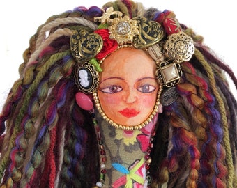 May include: A handcrafted doll with a painted face, adorned with a crown of vintage buttons and a beaded necklace. The doll's hair is made of colorful yarn in shades of green, red, and purple. The doll's face is framed by a fabric body.
