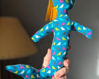 May include: A handmade mermaid doll with a blue body covered in colorful bird patterns. The doll has a yellow felt hair and a fishtail. The doll is being held up in front of a blurred background.