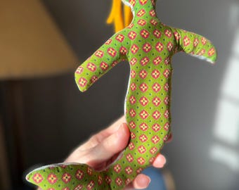 May include: A handmade mermaid doll with a green floral patterned body and yellow hair. The doll's body is covered in a repeating pattern of red and green flowers. The doll is soft and plush.