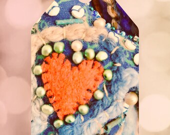 May include: A close-up of a handmade heart-shaped ornament with a red felt center, surrounded by blue and white fabric, beads, and twine. The ornament is decorated with a variety of beads and has a rustic, vintage aesthetic.