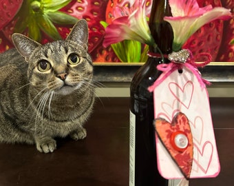 May include: A brown glass bottle with a pink and white flower attached to the neck. The bottle has a white label with a pink heart and the word "INCA" on the top. The bottle is sitting on a wooden table next to a tabby cat.