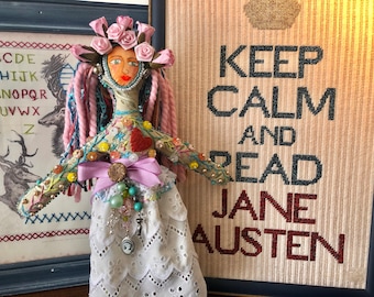 May include: A handmade doll with pink yarn hair, a floral dress, and a pink ribbon around her waist. She is wearing a white lace skirt and has a crown of pink roses on her head. The doll is standing in front of a framed cross-stitch that reads "Keep Calm and Read Jane Austen".