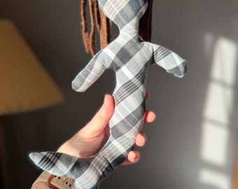 May include: A handmade mermaid doll with a plaid pattern in shades of gray and white. The doll has brown yarn hair and a fishtail. The doll is being held up in front of a gray background.
