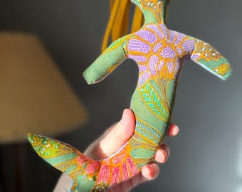 May include: A handmade mermaid doll with a green body and a patterned design in purple, orange, and yellow. The doll has a yellow hair and a fishtail. The doll is made of fabric and is being held by a person.