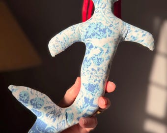 May include: A handmade mermaid doll with a blue and white patterned fabric body and tail. The doll has small arms and a red fabric hair piece. The doll is soft and plush.