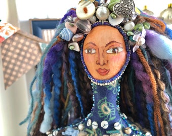 May include: A handcrafted doll with a painted face, adorned with a beaded necklace and a headpiece featuring shells, buttons, and beads. The doll has long, colorful yarn hair in shades of blue, purple, and brown. The doll's clothing is a dark blue fabric with a floral pattern.