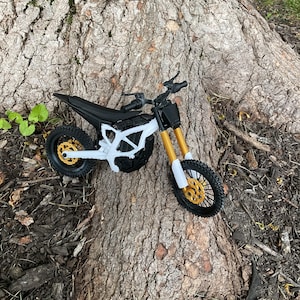 Könnte beinhalten: Ein Miniatur-Motocross-Modell mit weißem Rahmen, schwarzem Sitz und schwarzem Lenker. Die vorderen Federgabeln sind goldfarben, die Räder haben schwarze Reifen mit goldfarbenen Felgen und Bremsscheiben. Das Modell hat ein schwarzes Frontschild.