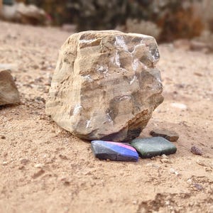 Puede incluir: Una gran roca marrón claro rugosa se encuentra sobre una superficie arenosa. Dos pequeños objetos rectangulares se encuentran delante de la roca. Uno es azul, rosa y negro, y el otro es verde oscuro.