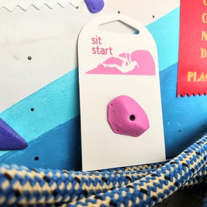 Op de afbeelding: Een roze klimgreep met een schroefgat, bevestigd aan een witte kaart met de tekst "sit start". De kaart toont ook een roze silhouet van een klimmer op een klimwand.