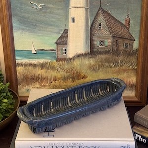 May include: An elongated, dark blue ceramic basket with a slatted, open-weave design and rope-textured handles at each end. The basket features a deep, rounded interior and a textured finish, resting on a stack of three hardcover books.
