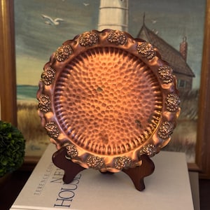May include: A round copper tray featuring a hammered center texture and a scalloped edge decorated with embossed floral motifs. The tray is displayed on a dark wooden stand, showcasing a warm metallic finish with subtle signs of natural patina.