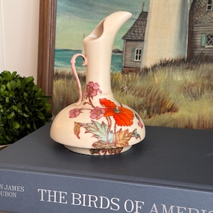 May include: A cream-colored ceramic pitcher with a narrow, angled spout and a small pink handle. The body features a painted floral design with a large, vibrant orange poppy and smaller pink flowers with green leaves on a smooth, rounded base.