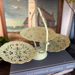 May include: A pale green metal tiered serving stand featuring three circular trays with intricate cut-out floral patterns and a central arched handle. The stand is constructed with a sturdy base and a vertical support rod connecting the three levels.