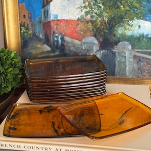 May include: A stack of rectangular amber-colored glass plates with dark, swirling marbled patterns. Two additional plates are positioned in front, displaying the same translucent, warm-toned glass and distinct dark streaks throughout the surface.
