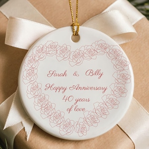 Op de afbeelding: Een witte keramische ornament met een gouden touwtje, met een hartvormig rozenontwerp. Het ornament heeft de tekst "Sarah & Billy Happy Anniversary 40 years of love" in rode letters. Het ligt op een ingepakt cadeau met een crème lint.