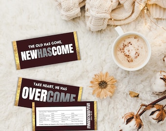 May include: Two chocolate bar wrappers with gold foil accents. The top wrapper reads "The old has gone, New has come". The bottom wrapper reads "Take heart, he has overcome".