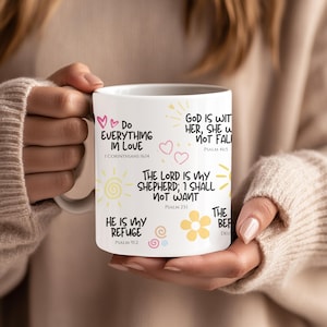 May include: White ceramic mug featuring inspirational quotes and Bible verses in black script, with pink and yellow decorative elements. The mug includes phrases like "Do Everything in Love" and "The Lord is My Shepherd."