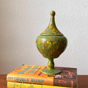 Puede incluir: Un jarrón verde de cerámica con tapa y detalles en naranja y amarillo se encuentra sobre una pila de libros antiguos. Los libros tienen cubiertas naranjas y amarillas con títulos como "The Double Blind". El jarrón tiene una base de pedestal.
