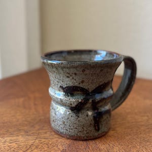 May include: A ceramic mug with a speckled grey and brown glaze, featuring a flared rim, a central horizontal ridge, and a dark brown abstract brushstroke pattern. The mug has a sturdy handle and a visible reddish-brown clay base at the bottom.
