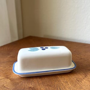 May include: A rectangular white ceramic butter dish with a lid. The lid features a hand-painted design of two light blue leaves flanking three small dark blue berries. The base of the dish has a thin blue rim around the edge of the tray.