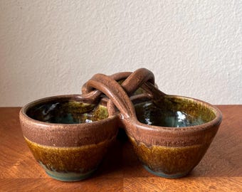 May include: A double-bowl ceramic server featuring two connected round vessels joined by a central arched handle. The exterior displays a two-tone glaze with a dark brown upper rim and a mottled golden-brown base, while the interior is green.