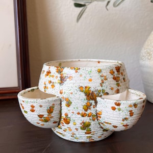 May include: A ceramic strawberry planter with a speckled off-white glaze featuring orange and green paint splatters. The vessel has a wide central opening and two smaller side pockets, all with a ribbed texture along the exterior surface.
