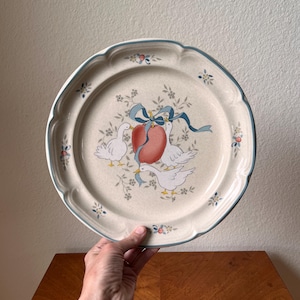 Puede incluir: Un plato de cerámica beige con borde festoneado y ribete azul. El plato presenta un diseño de tres gansos blancos, una manzana roja y cintas azules. Acentos florales se dispersan alrededor del borde.