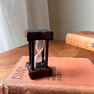 Puede incluir: Un pequeño reloj de arena de madera oscura con arena rosa, colocado sobre un libro antiguo. El reloj de arena tiene un diseño clásico, con una parte superior e inferior rectangulares. El libro es de color naranja apagado y tiene el título "Riley Love".