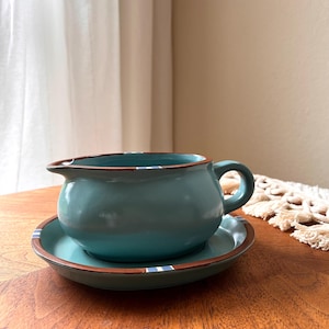 Può includere: Un set di salsiera e piattino color turchese. La salsiera ha un manico e un beccuccio, e il set presenta un bordo marrone con strisce blu e bianche. Il set è su una superficie di legno.