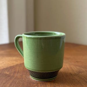 May include: A ceramic mug featuring a light green glazed upper section with a visible crackle finish. The base of the mug is a contrasting dark brown band decorated with a repeating pattern of small, light-colored dots and a thin white line.