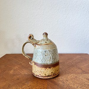 May include: A handcrafted ceramic mug with a lid. The mug features a textured, light blue upper section, a brown band, and a yellow-brown base. It has a curved handle and a small, rounded lid with a knob.