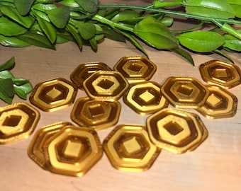 May include: A collection of gold-colored, hexagonal tokens with a diamond-shaped center. The tokens are arranged on a light-colored wooden surface, with green leafy sprigs in the background. The tokens have a metallic sheen.