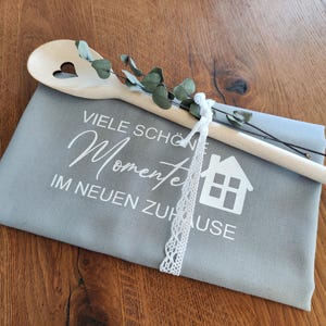 Könnte beinhalten: Ein Holzlöffel mit herzförmigem Ausschnitt liegt auf einem grauen Stoff mit weißem Text und einer Hausgrafik. Grünzeug und ein weißes Spitzenband sind enthalten. Der Text lautet "Viele schöne Momente im neuen Zuhause."