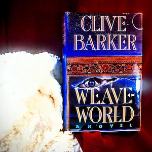 May include: A hardcover book titled "WEAVEWORLD" by Clive Barker. The book has a blue cover with gold lettering and an eye graphic. The book is next to a white, textured object on a red surface.