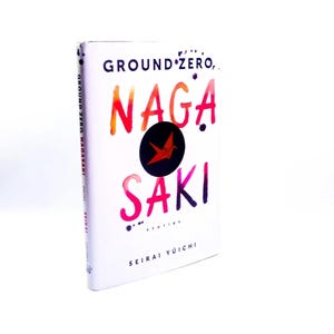 May include: A white book titled "GROUND ZERO, NAGASAKI" with colorful text. The cover features a black circle with a red bird graphic. The author's name, Seirai Yoichi, is at the bottom. The book's spine also has the title.
