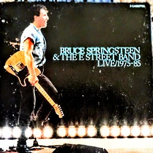 May include: Album cover for Bruce Springsteen & The E Street Band's "Live/1975-85." The image features a man holding a guitar, with stage lights in the background. The album title and artist name are in a teal font against a dark background.
