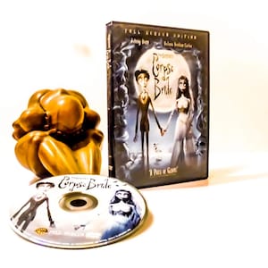 May include: A DVD case and disc for the movie "Corpse Bride" featuring Johnny Depp. The case shows a full moon and the characters Victor and Emily. A golden-brown sculpture is also in the image.
