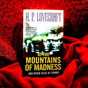 May include: A book titled "At the Mountains of Madness" by H.P. Lovecraft. The book cover features the title in yellow and the author's name in white. The book is set against a red, textured background.